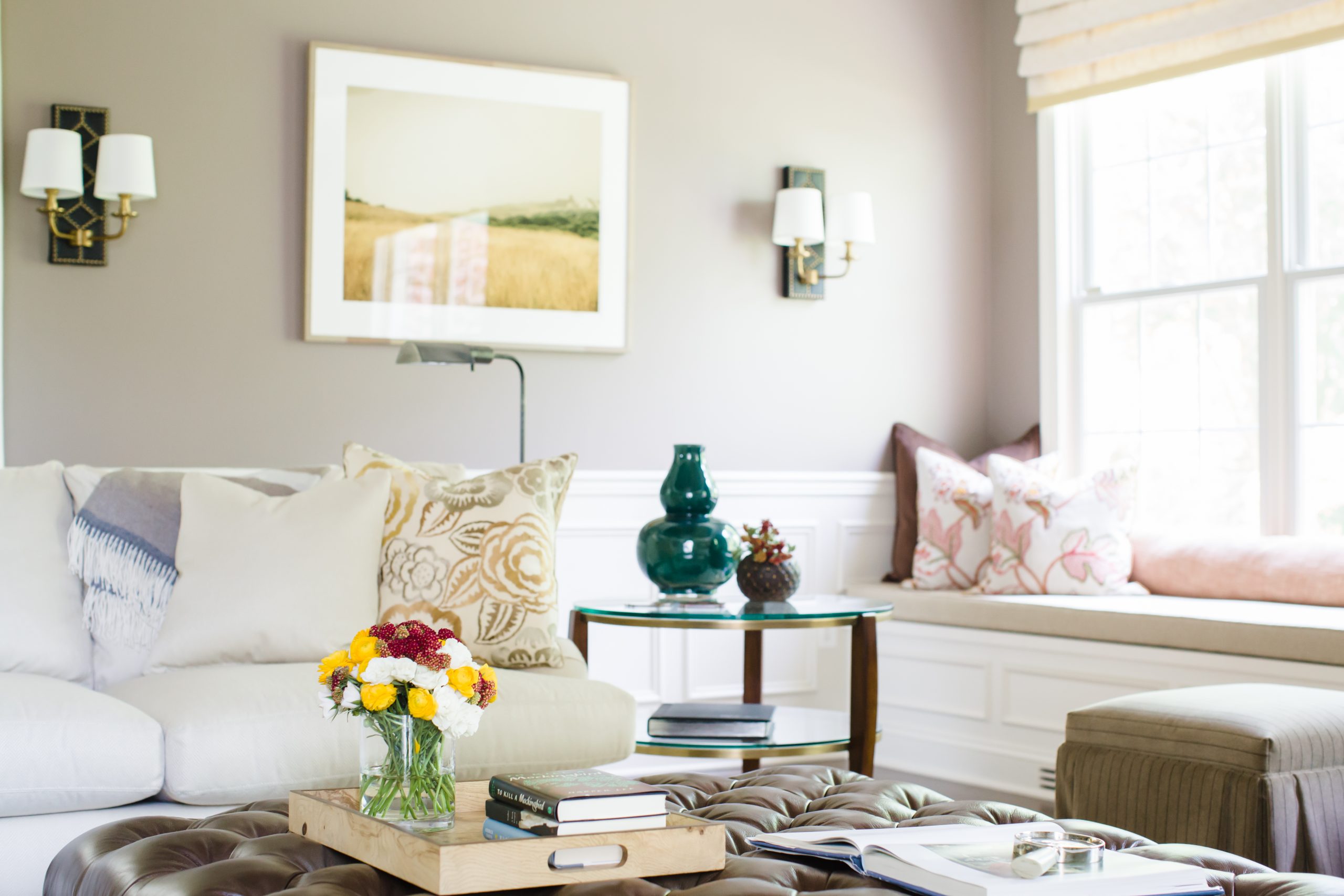 Transitional family room with decorative white molding on walls, grey wall color, glass side table, beige furniture cushions, and a brown leather tufted ottoman