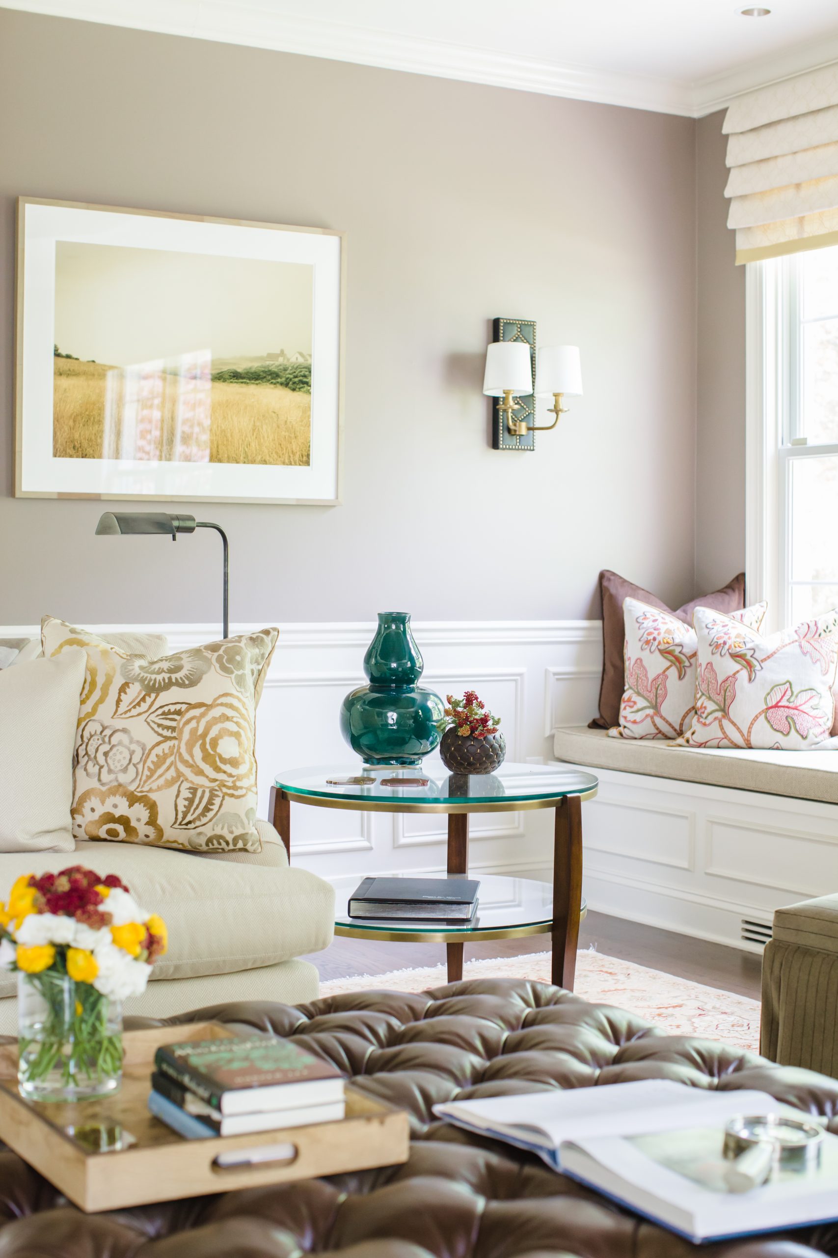 Transitional family room with decorative white molding on walls, grey wall color, glass side table, beige furniture cushions, and a brown leather tufted ottoman