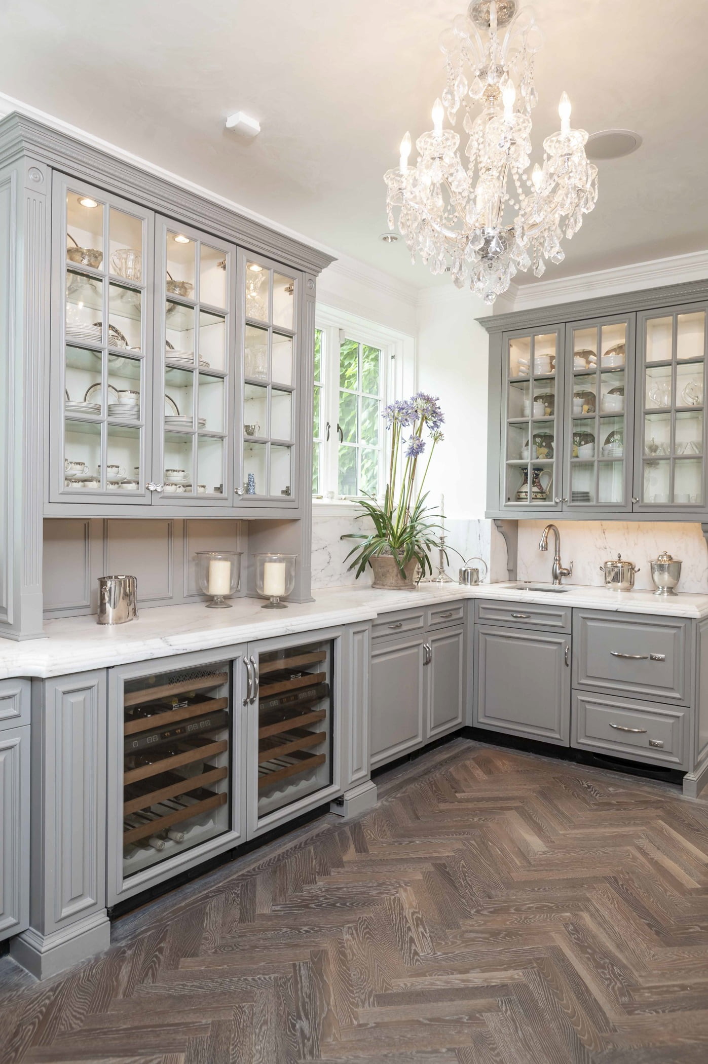 Traditional kitchen area with custom light grey cabinetry, marble countertops, ornate crystal chandelier, and dark chevron hardwood flooring (Different view)