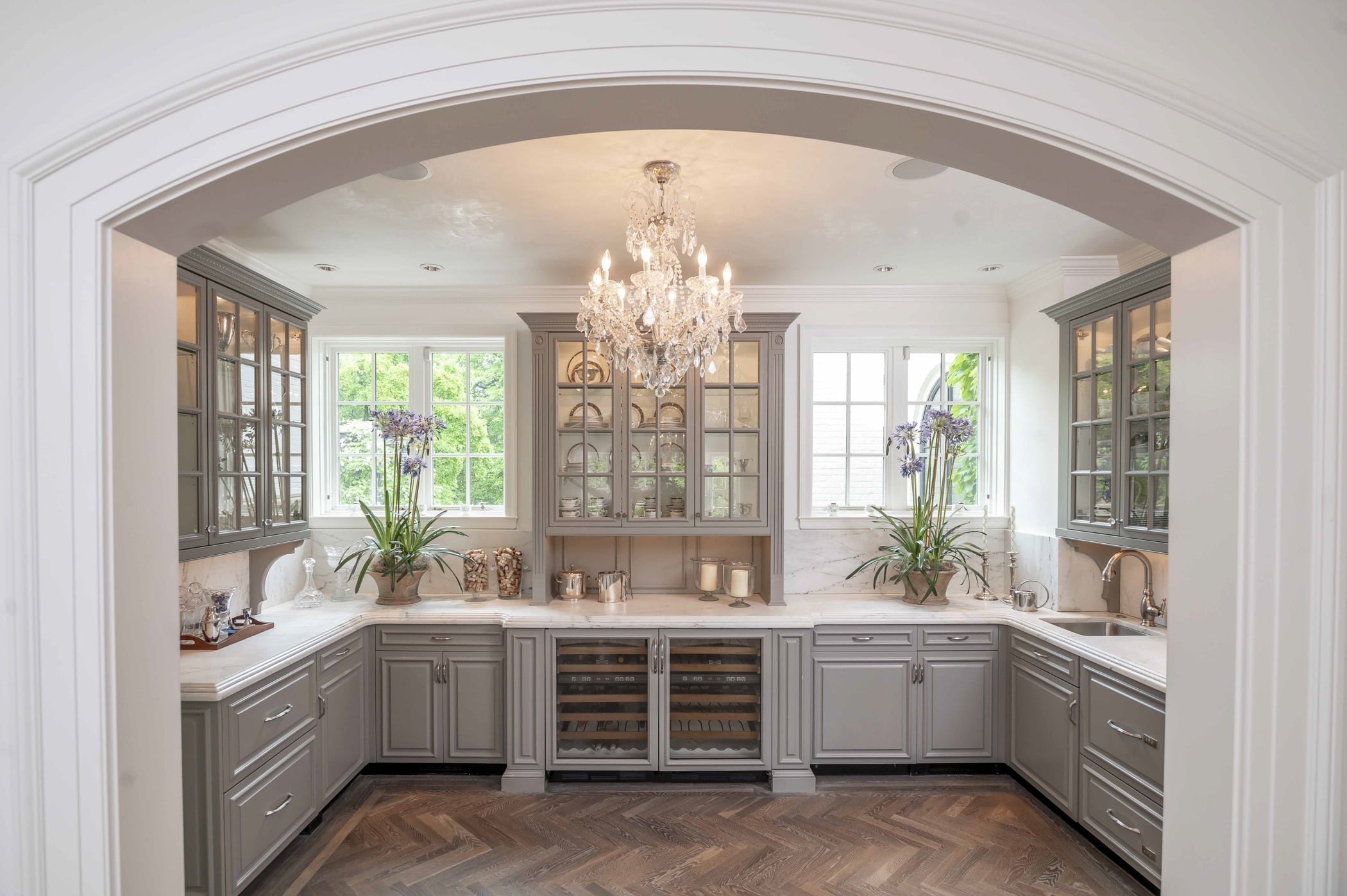 Traditional kitchen area with custom light grey cabinetry, marble countertops, ornate crystal chandelier, and dark chevron hardwood flooring