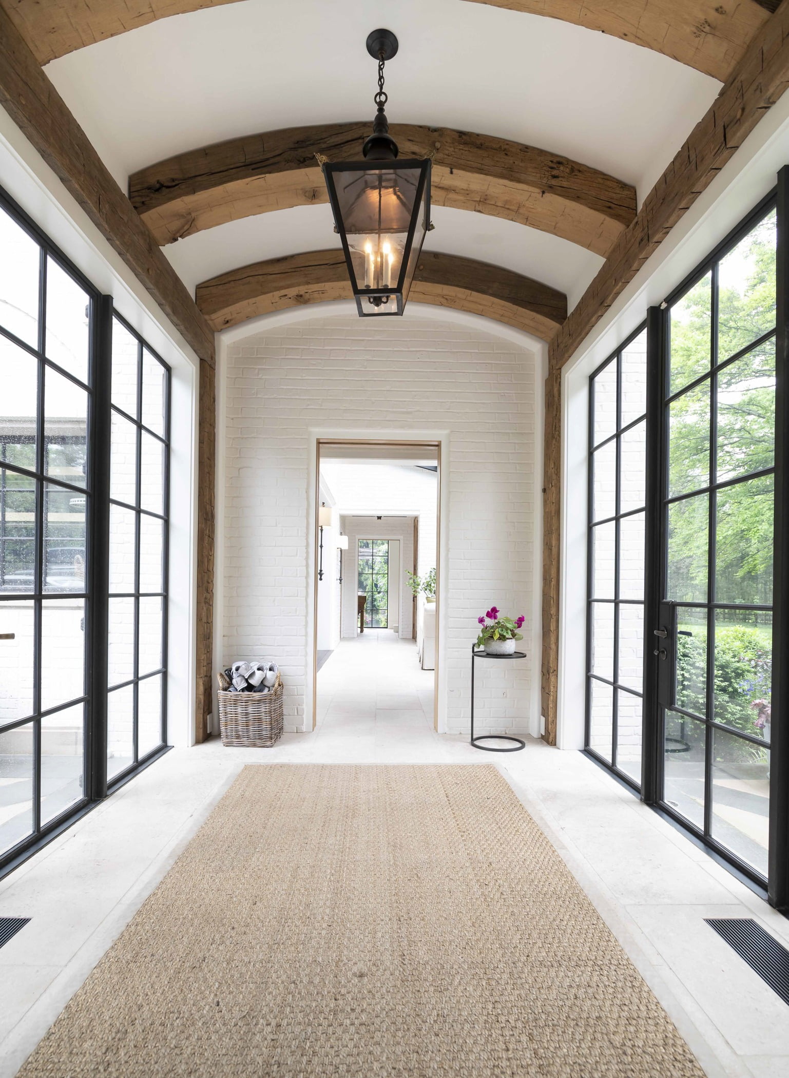 Contemporary/modern hallway with thick wooden ceiling arches, white tile flooring, and black steel framed doors and windows on either side (3)