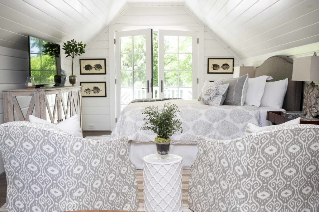 Transitional bedroom with white panel walls and ceiling, light grey accent color with bed and armchairs, and dark hardwood flooring with beige carpet